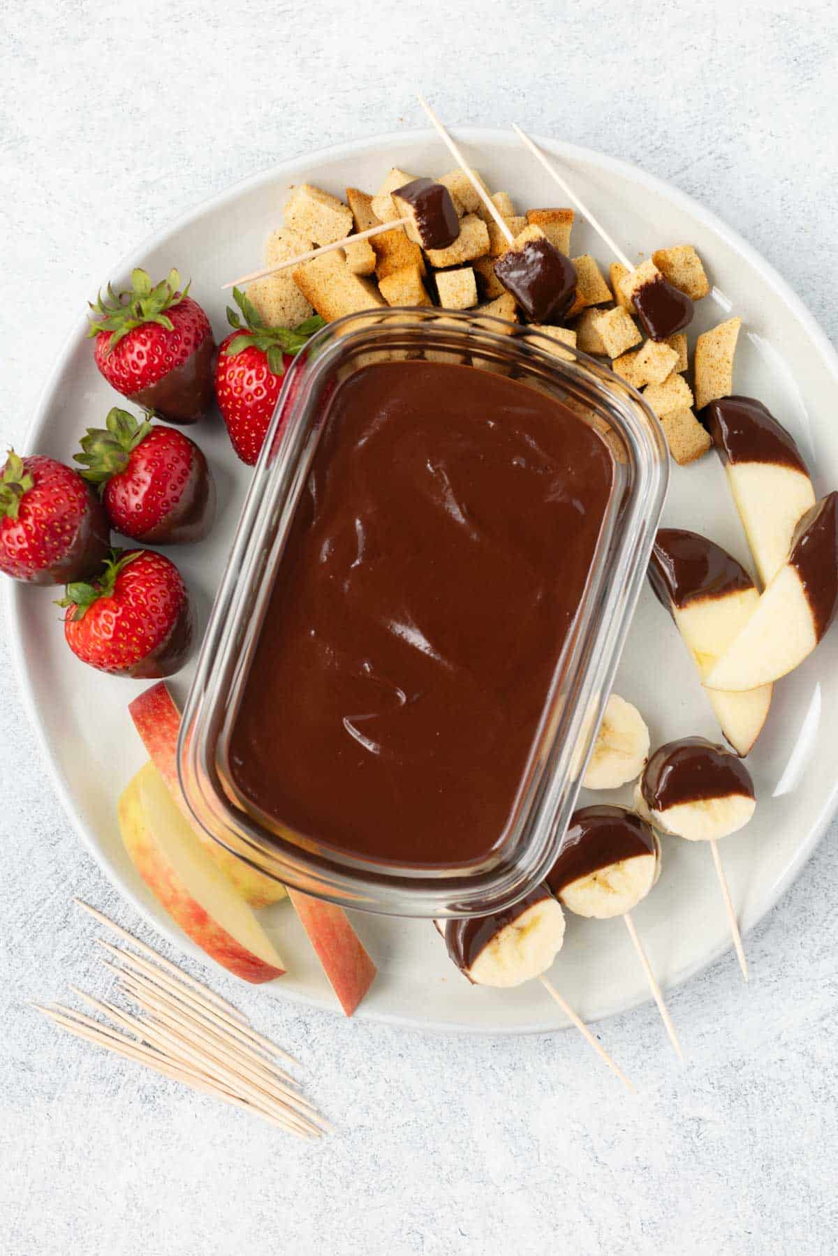 A container of homemade chocolate sauce on a plate surrounded by fruit and bread cubes.