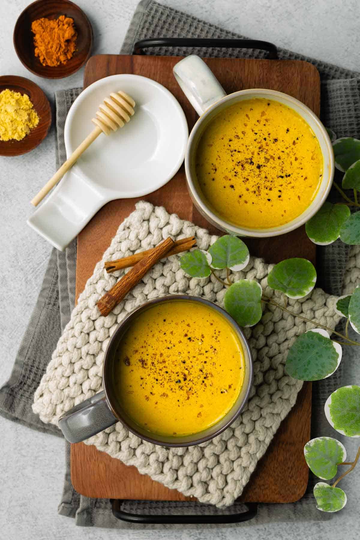 Two mugs of golden milk are resting on a light gray rope trivet with a walnut wood tray underneath.