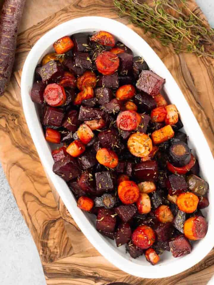 Roasted beets and carrots in a white oval serving dish, resting on a rustic wood cutting board.