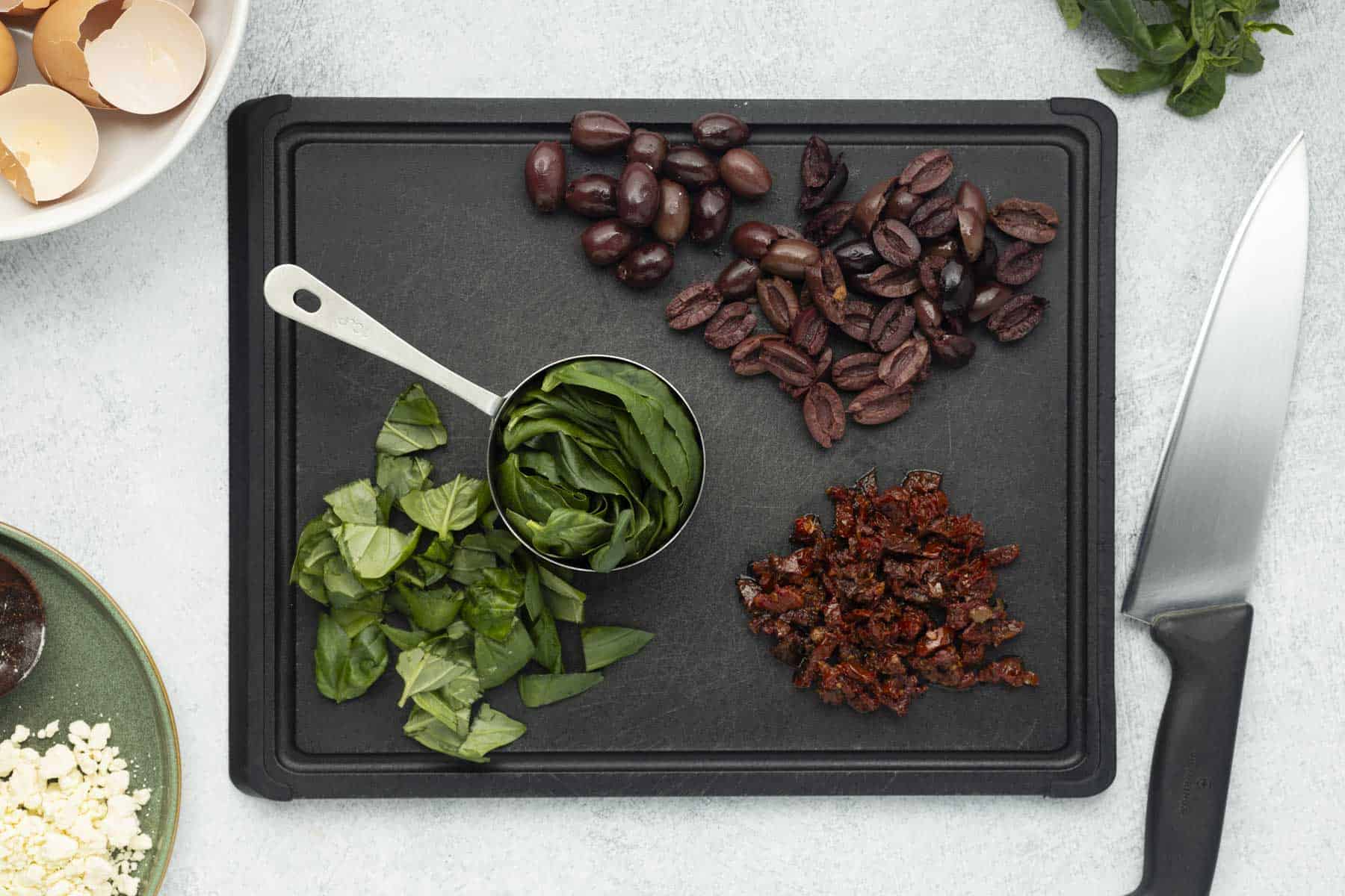 Chopped kalamata olives, sundried tomatoes, and basil on a cutting board, next to a chef's knife.