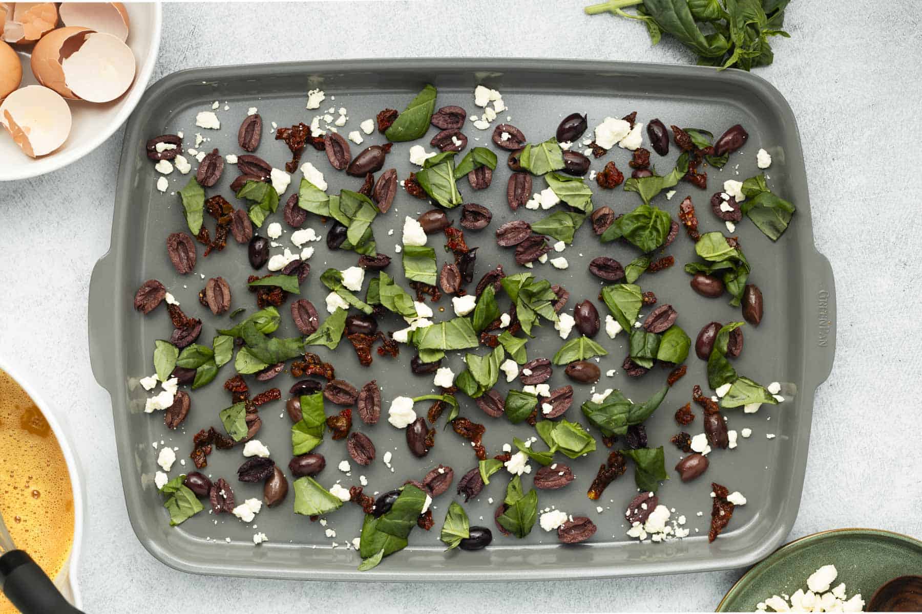 Chopped olives, sundried tomatoes, and basil evenly spread on a baking sheet with crumbled feta cheese.