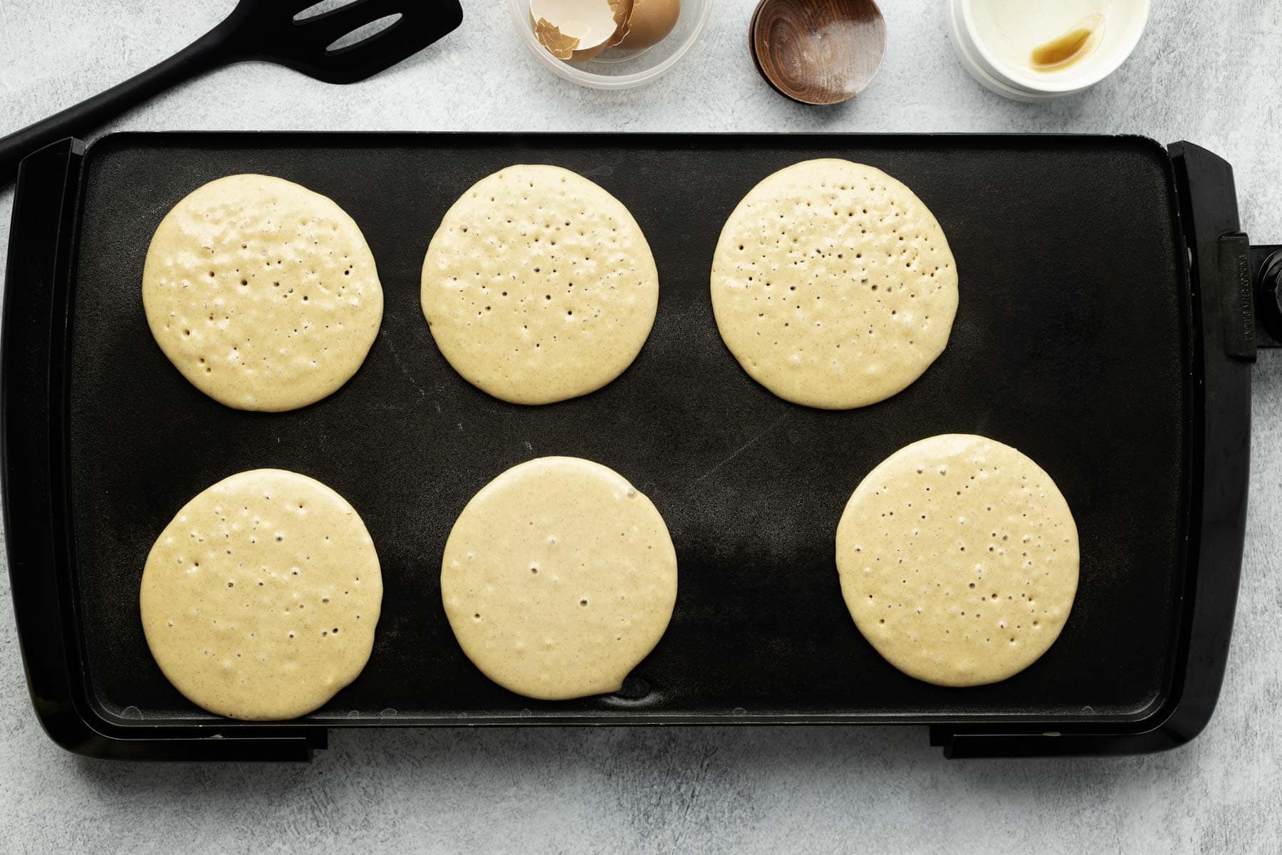 Six pancakes cooking on a black griddle, not yet flipped.