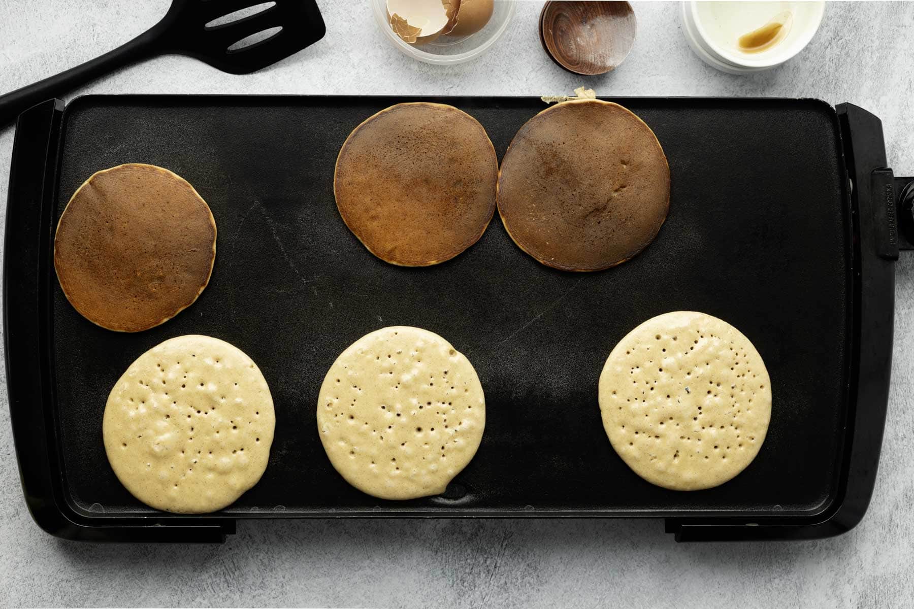 Six pancakes cooking on a black griddle with three flipped and three not yet flipped.